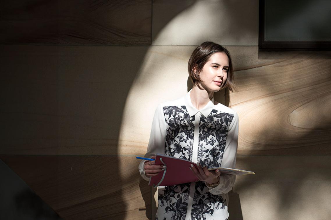 Women with book stands in speckled light against sandstone wall.